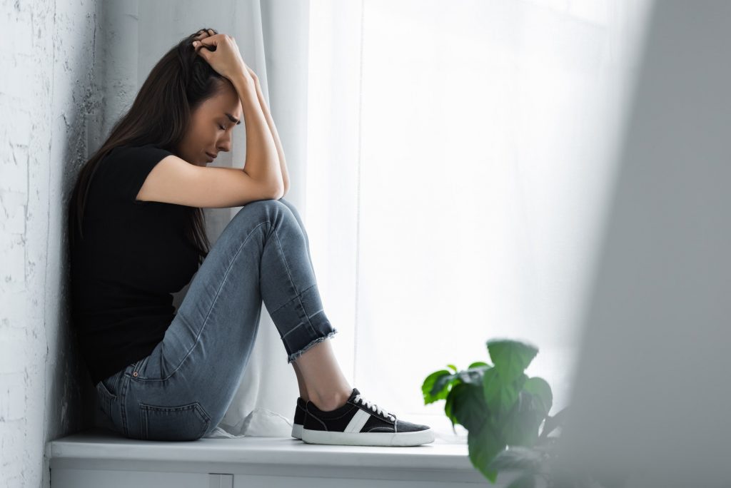 selective focus of young woman crying while sitting on window sill and suffering from depression